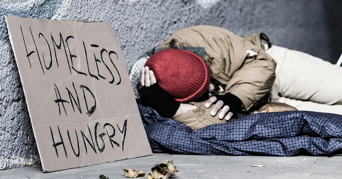 Photo of a homeless person sleeping outdoors on a sidewalk, next to a sign that says "homeless and hungry." Photo by Photographee.eu on Adobe Stock.