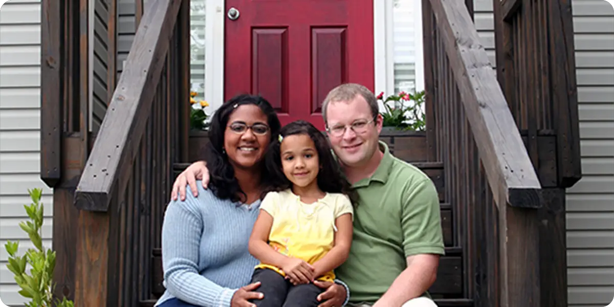 A happy family of three is seated together on the porch of their new home. A woman in a blue sweater on the left, and a man in a green shirt on the right sit with a young girl in yellow between them.