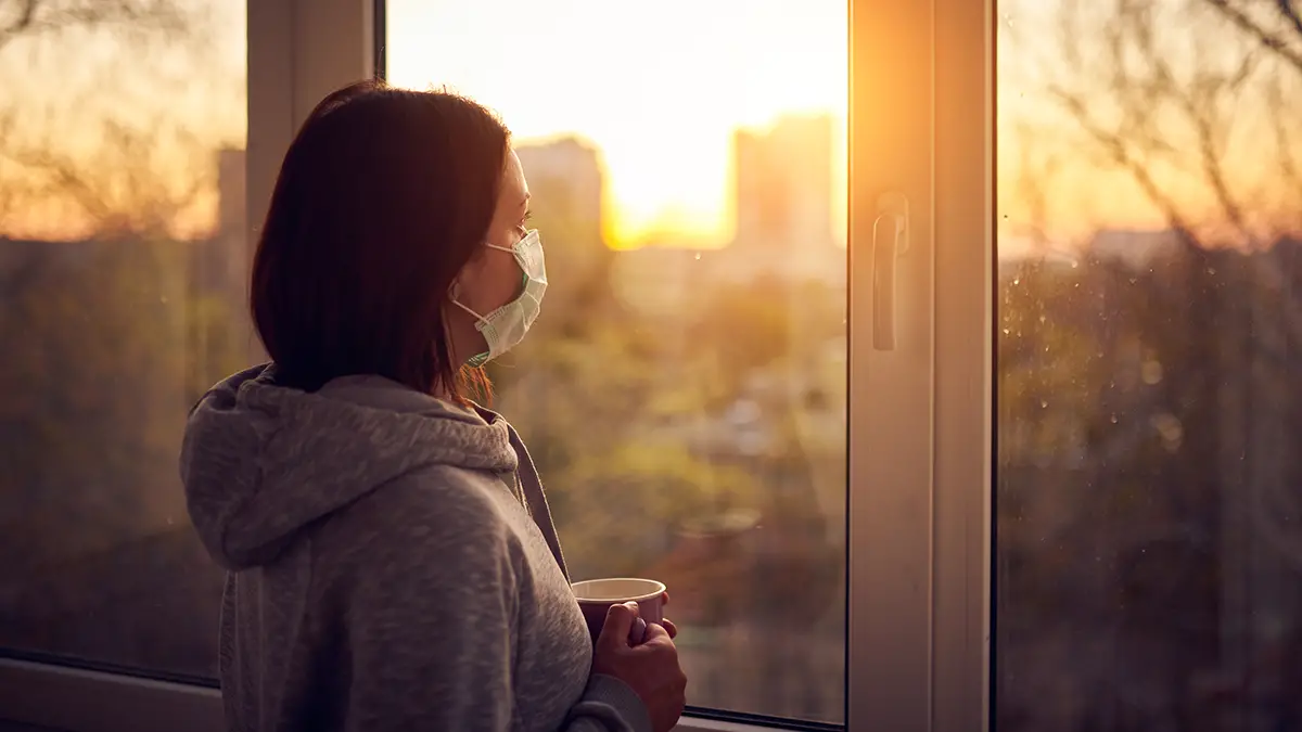 Photo of a woman with a medical mask over her face, and looking out at a sunrise through a window.
