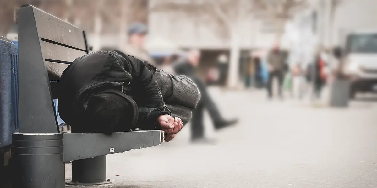 Photo of a homeless person laying down on a bench during the day that's located on a busy city street.