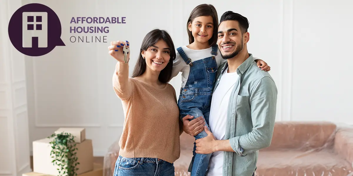 Photo of a family of a mom, dad and child smiling while standing in a living room while the mom holds up house keys, with the Affordable Housing Online logo.